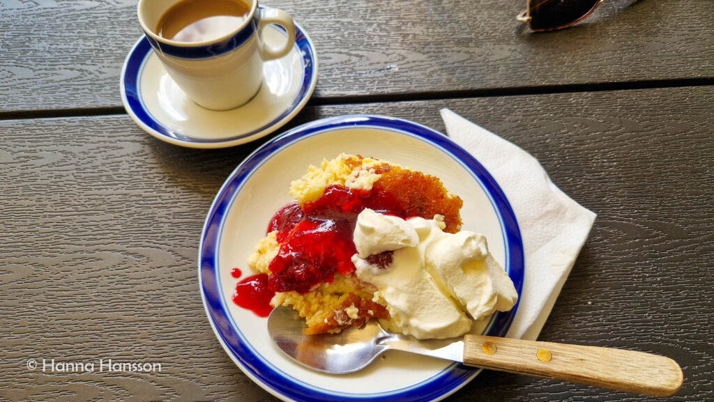 En assiett med ostkaka, körsbärssylt och grädde bredvid en kopp kaffe.