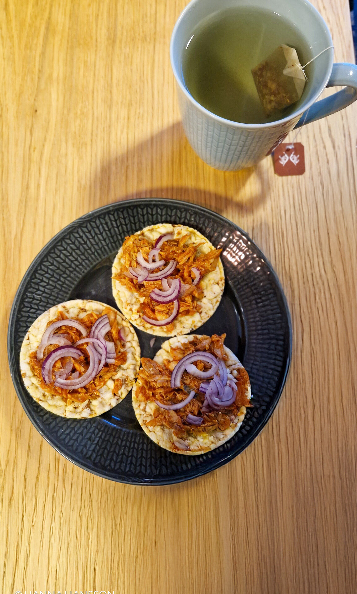 Tre riskakor toppade med makrill i tomatsås och skivad rödlök, serverade på en svart tallrik med en kopp grönt te på ett träbord. Ett enkelt och nyttigt mellanmål med omega-3 och protein.