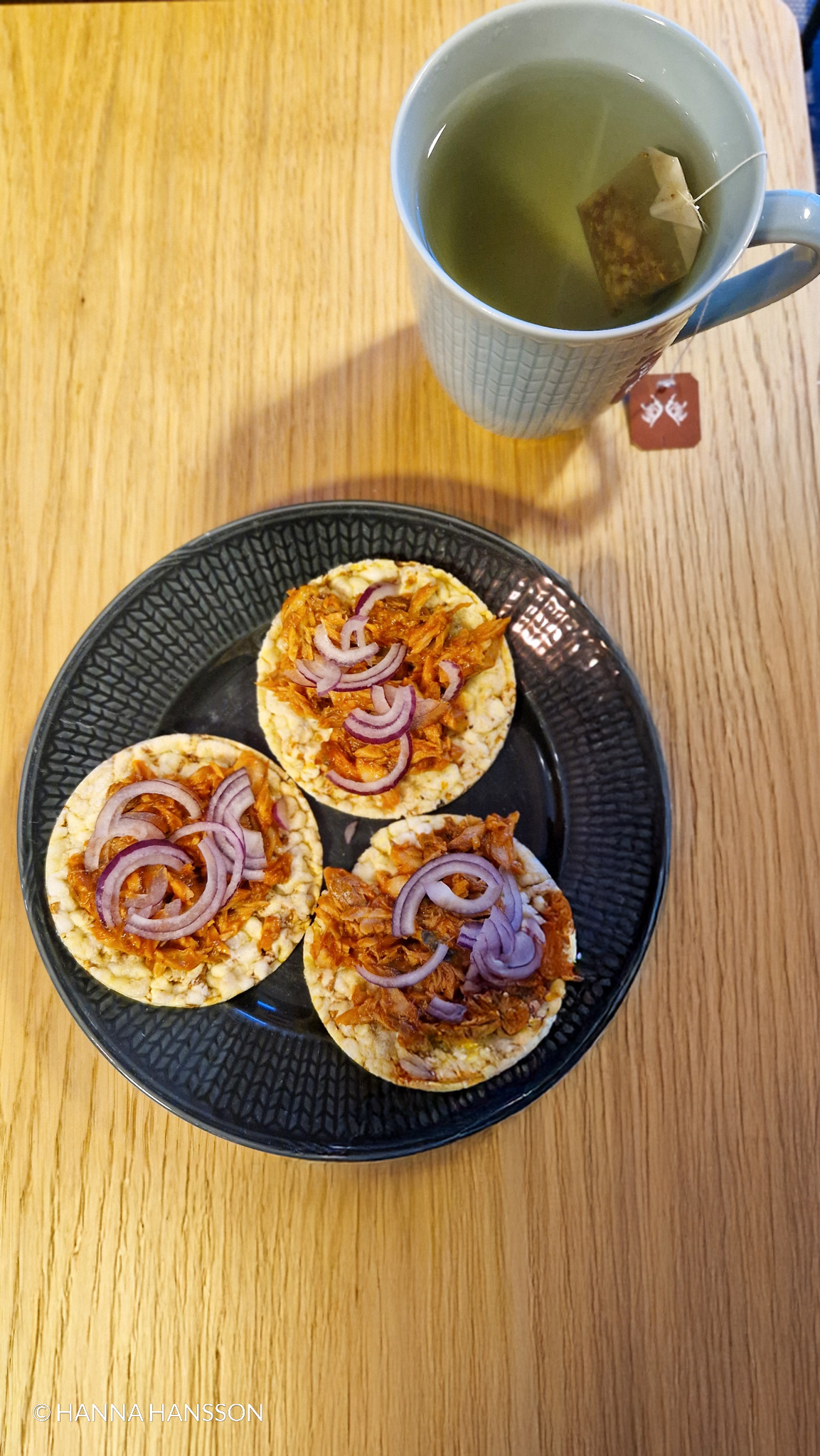 Tre riskakor toppade med makrill i tomatsås och skivad rödlök, serverade på en svart tallrik med en kopp grönt te på ett träbord. Ett enkelt och nyttigt mellanmål med omega-3 och protein.